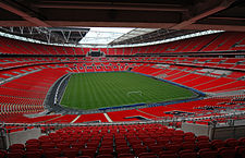 The interior of an empty stadium as viewed from its upper tier of seating. The seats are a vivid red and the pitch is a vivid green. The pale grey sky is visible through an opening in the ceiling above the pitch