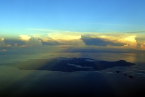 The sun sets behind the clouds, on the horizon over a small island.