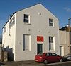 Three-quarter view of a tall, two-storey, plain-walled, white-painted building with a pointed roof. The front has one blank window and a half-blank, half-glazed window flanking a narrow door, above which is an orange sign.  There are also six circular protrusions.  The side wall has three windows and a thin metal flue.