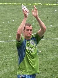 The head and torso of a young man, wearing a green and blue top and blue shorts, standing on a grass field. He has his arms raised above his head in what appears to be a clapping motion.