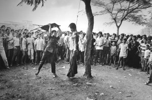 Thammasat University massacre: A crowd looks on, some with smiles on their faces, as a man uses a folding chair to beat the hanged body of a student just outside the university