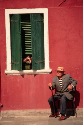 An elderly resident of Burano sits in the sun outside his home, dressed nattily in a straw hat and jacket.