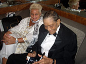 A man in a suit and a woman in a white outfit seated on a couch; both are looking toward reading material he is holding in his hands along with a pen.