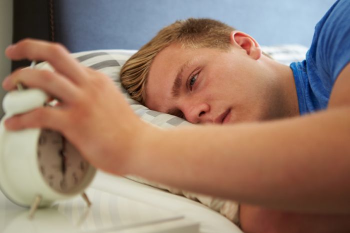 A sleepy teen with his hand on an alarm clock