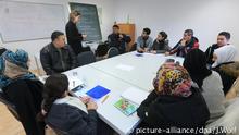 Eine Gruppe von syrischen Deutschlernern sitzt um einen Tisch herum. eine Lehrerin steht an der Tafel. 