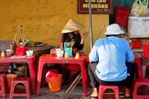 A woman selling Cau Lau, a local noodle specialty on a street sidewalk in Hoi An, Vietnam.