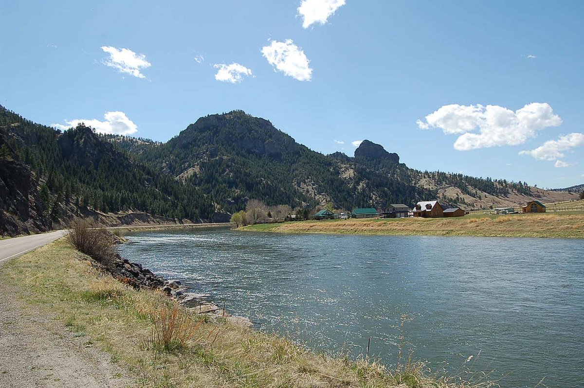 Tree covered hills and a river surround a road that leads to a cluster of old buildings. The sky is light blue with a few scattered white clouds.  Old U.S. Highway 91 Historic District, Montana