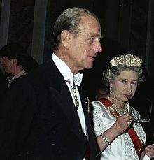 Elizabeth, in formal dress, holds a pair of spectacles to her mouth in a thoughtful pose