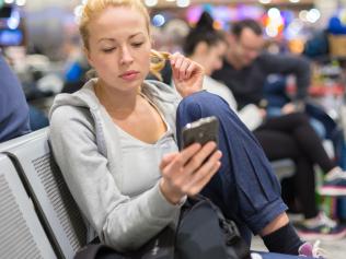 Casual blond young woman using her cell phone while waiting to board a plane at the departure gates. Wireless network hotspot enabling people to access internet conection. Public transport.