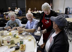 In this Thursday, May 7, 2015 photo, volunteer Sandy Lockhart, standing, serves lunch to John Pearson, right, as Dorothy Christian, left, and her husband, Fredrick, look on at a seniors meal program at the Rush Park