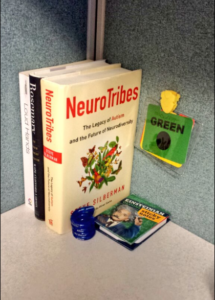 Three books are in the corner of a cubicle. The titles are "Neurotribes," "Rosemary," and "Loud Hands." Beside them, a color communication badge is attached to the cubicle wall. A stim toy and Einstein sticky notes are holding the books in place. 