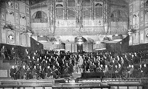 large orchestra and their conductor seen on the platform of Victorian concert hall in long shot