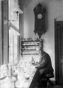 An old, bespectacled man wearing a suit and sitting at a bench by a large window. The bench is covered with small bottles and test tubes. On the wall behind him is a large old-fashioned clock below which are four small enclosed shelves on which sit many neatly labelled bottles.