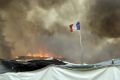 A French flag flutters while smoke billows from burning shelters set on fire by migrants.