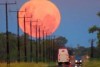 A red full moon rises over vehicles driving along a rural road, with powerlines beside the road disappearing into the distance.