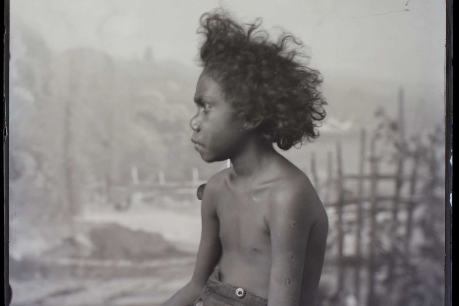 An Aboriginal child sits on a stool in a studio waiting to have his portrait photographed