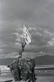 Israeli soldiers stabilize a flag pole whilst another soldier climbs it in order to raise an improvised flag; the soldier is seen about halfway up the flag pole. Other soldiers look on.