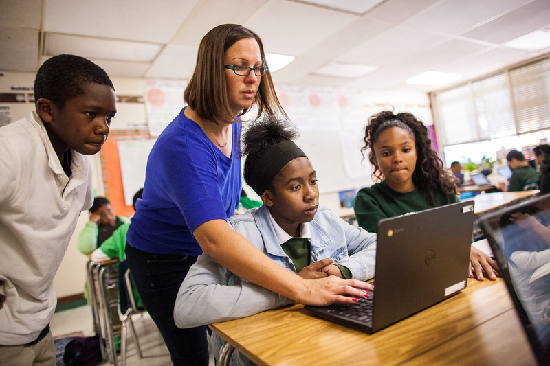 A teacher helps a student on a laptop, with two other students watching