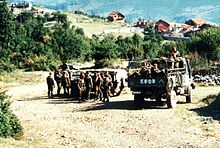 Three trucks of soldiers idle on a country road in front of trees and red roofed houses. The rear truck has KFOR painted on is back.