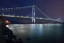 A tall suspension bridge connects a distant piece of land at night.