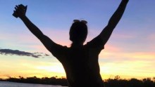 The dark silhouette of a woman with her hands in the air in front of the Pelican Point Billabong at sunset.