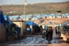 Two men ride a motorbike through a refugee camp of makeshift brick houses with blue tarpaulin roofs