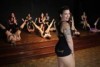 A woman with long hair and tattoos stands in front of a group of burlesque performers who are wearing training costumes