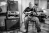 A man sits next to a series of amplifiers and speakers, holding a guitar in his hands.