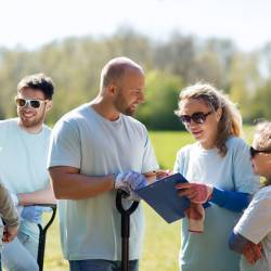 A group of people in matching teeshirts in a park, smiling, talking, and looking at a clipboard.