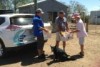 Three people and a dog surround the boot of a car as donated grocery items are handed over