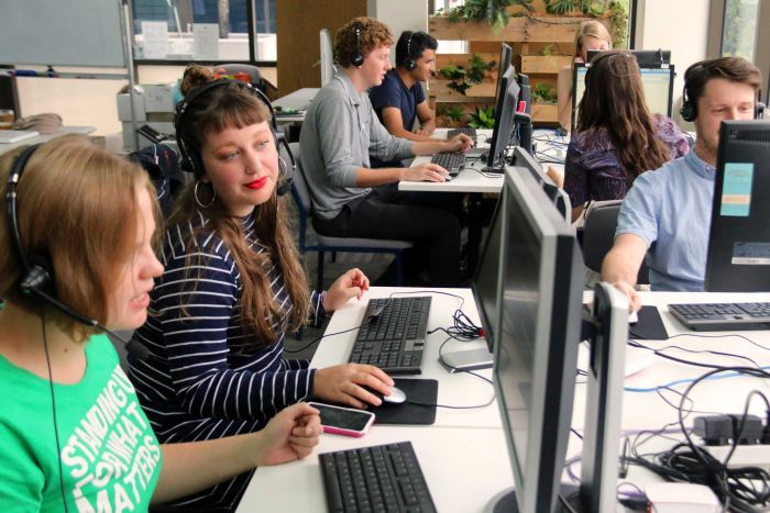 A group of people sit at desks and computers with telephone headsets on in an office.