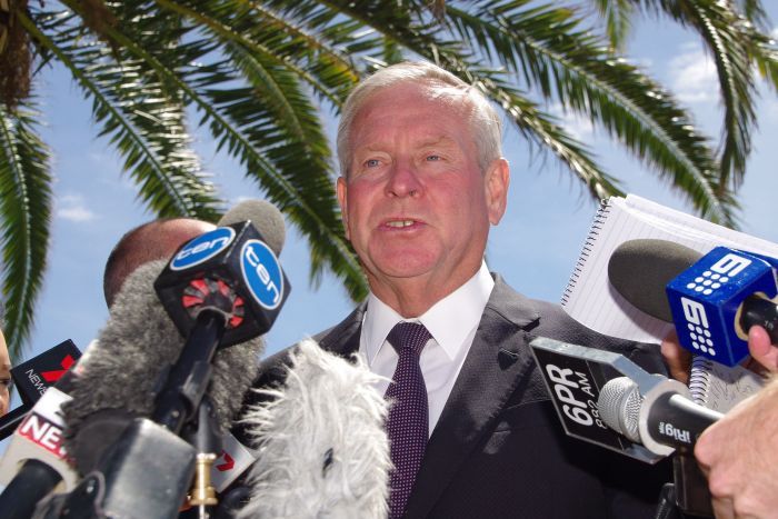 Upshot of Colin Barnett surrounded by microphones under a palm tree.