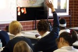 A girl raises her hand in a high school classroom surrounded by classmates.