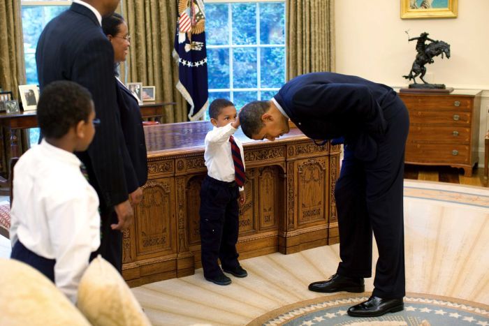 Barack Obama bends over so that the son of a White House staff member can pat his head.