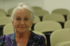 An old lady with grey hair is smiling in a museum surrounded by chairs. 