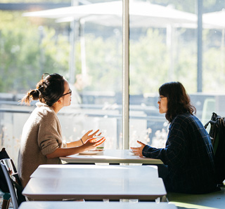 Two women having a conversation at a table near a large window.