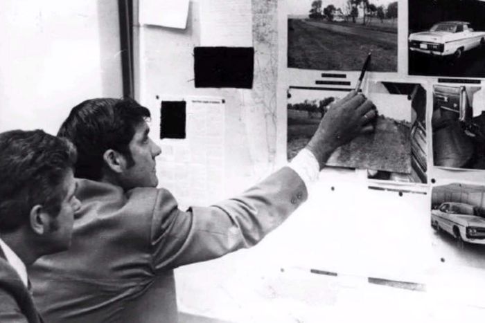 A black and white photo showing the back of two men pointing at an investigation pinboard