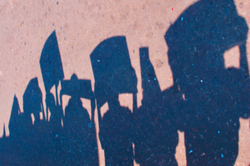 File: The silhouettes of activists holding signs seen in shadow on pavement. (Modified from Flickr / Vikalpa)