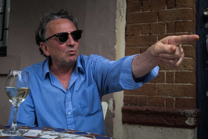 Playwright Louis Nowra sits at a table, pointing, mid-conversation