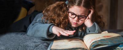 A girl lay on top of a bed with a large old book and hovers her hand over the page as she reads, (Maud).