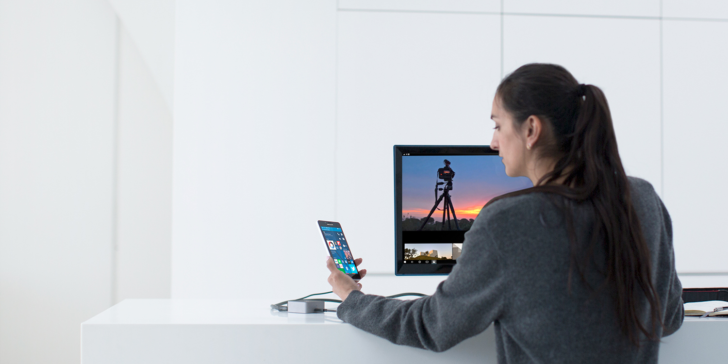 Woman sitting at desk, looking at her Lumia phone with Windows 10 start screen