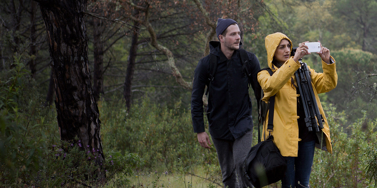 Woman and man wearing rain gear walking through rainy forest, the woman is carrying camera gear and takes a photo with her white Lumia 950