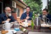 Three men sit around a table laughing while they share morning tea.