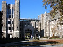 Four-story tower on left beset by arched walkway in center and pedestrian bridge connecting tower to three-story Gothic building