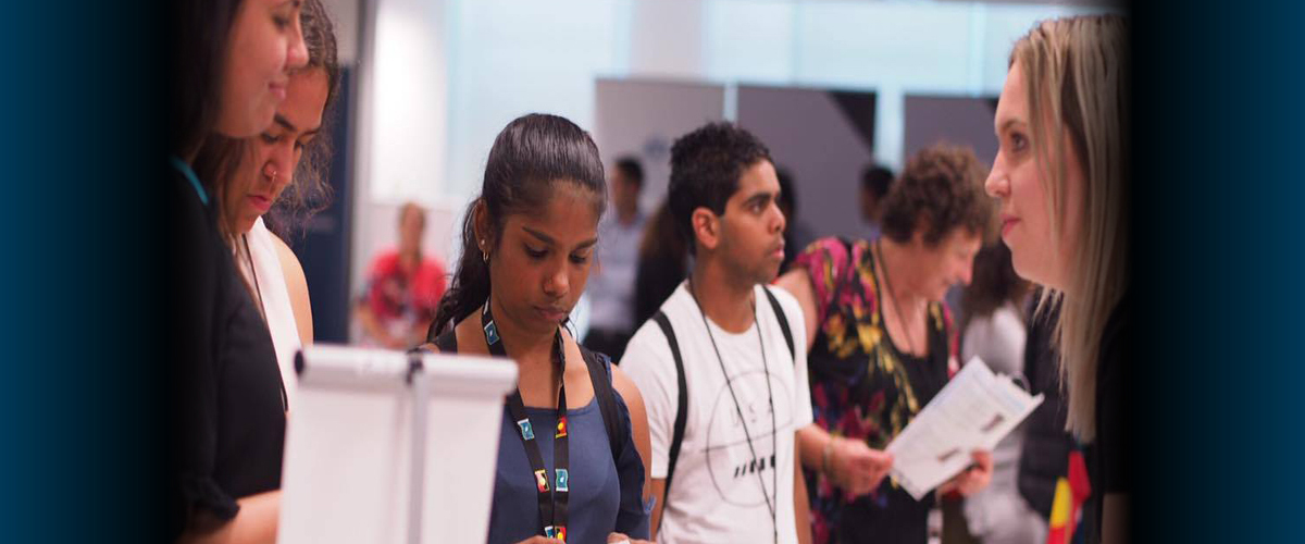 Four Indigenous young adults stand in front of a stall receiving advice from a non-Indigenous woman with other people standing in the background.