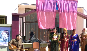 Women stand on a platform at an event while a man plays oud