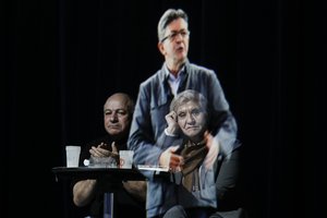 People sit at a table on stage and look towards the hologram of hard-left French presidential candidate Jean-Luc Melenchon, as he speaks to supporters who are gathered in Saint-Denis, near Paris