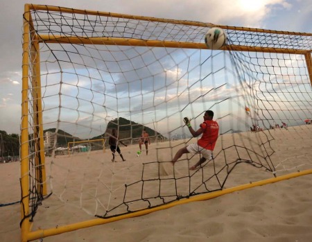 A goalkeeper misses a penalty kick at sunset in the sand on Copacabana Beach.
