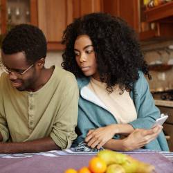 A man looking down at his phone; a woman next to him, looking over his shoulder at the phone, suspicious