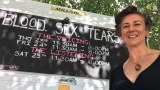 Woman standing in front of caravan with blackboard reading "blood, sex, tears".